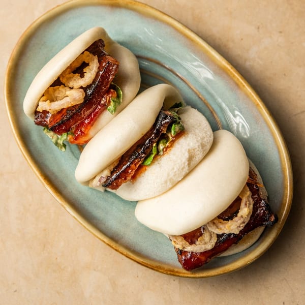 Three AquaSan Asian bao buns with glazed pork belly and crispy onions on a blue plate.