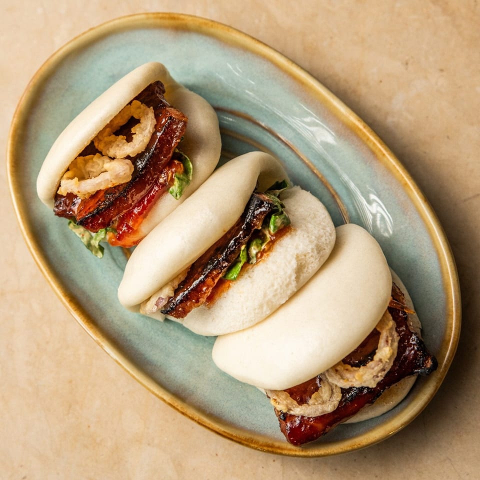 Three AquaSan bao buns with glazed pork belly, crispy onions, and greens on a blue plate.