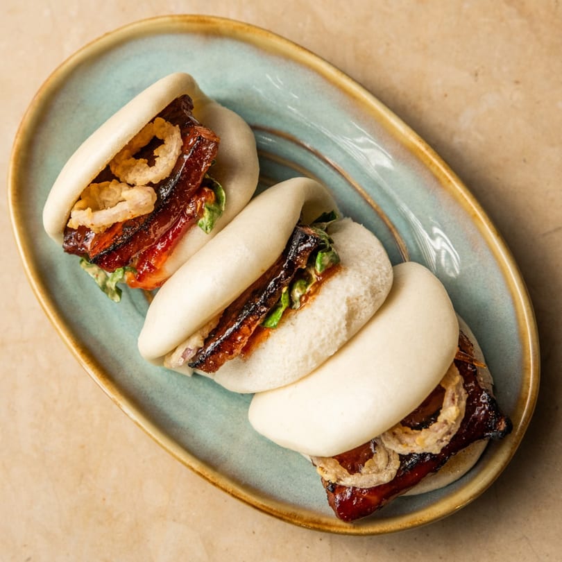 Three AquaSan bao buns with glazed pork belly, crispy onions, and greens on a blue plate.