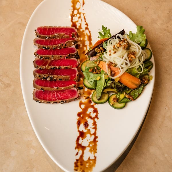 Seared tuna slices with sesame seeds and a side salad with noodles and vegetables on a white plate.