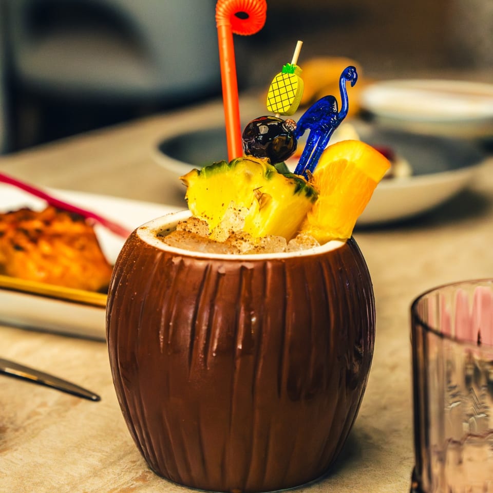 A tropical cocktail in a coconut-shaped glass, garnished with pineapple, orange, and a flamingo pick.