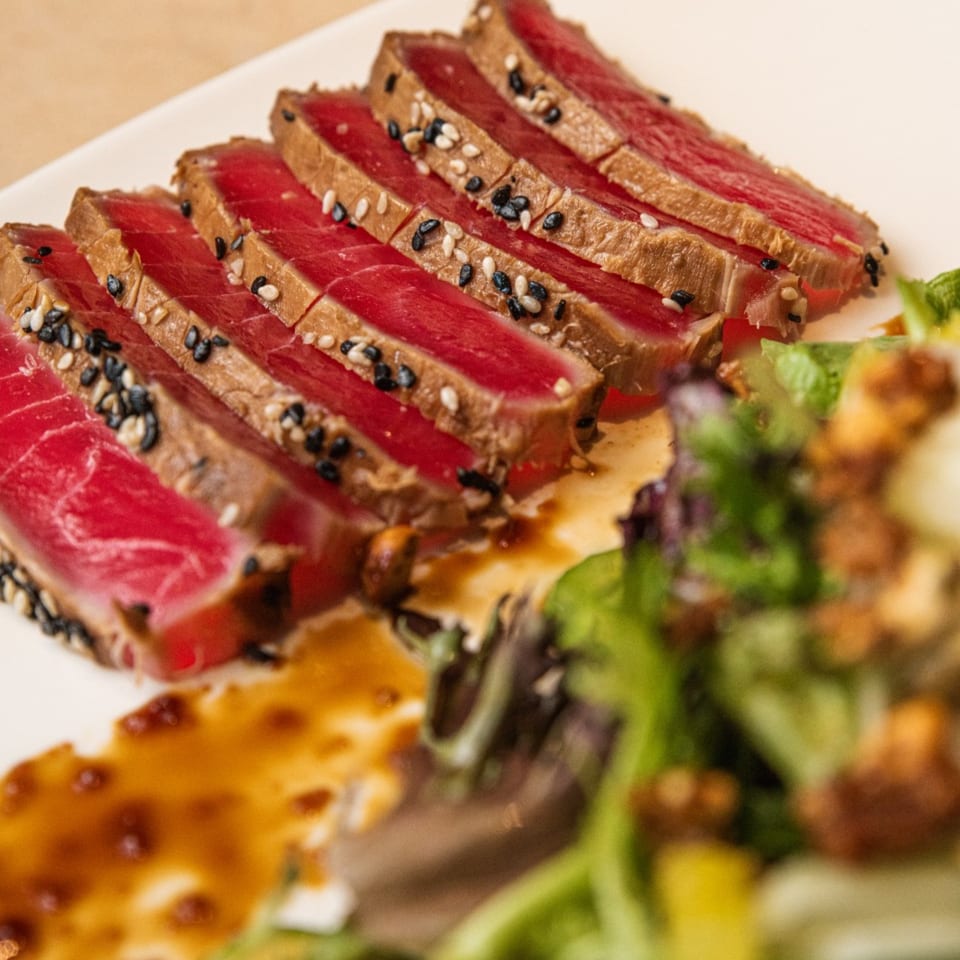 Seared tuna slices with sesame seeds, drizzled with sauce, and a side of fresh salad.